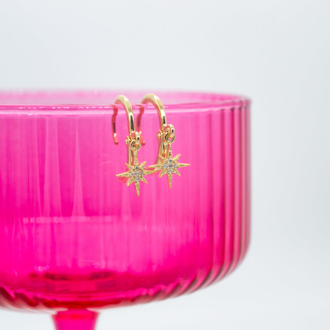 Gold earrings with star designs hanging from a pink container on a white background